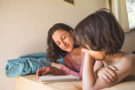 A boy with his mother lies in bed and reads a book, A woman spends time with son at home, A lazy weekend with a child, Portrait of a toddler with mother, Mom entertains the child during quarantine.の写真素材