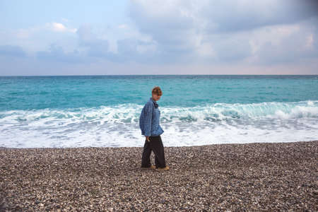 A girl with short red hair is walking along the beach, a hipster, a woman is walking along the sea, a girl in a long shirt and wide pantsの写真素材