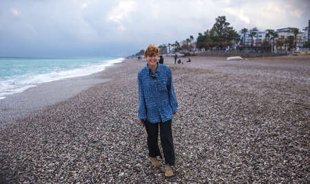 A girl with short red hair is walking along the beach, a hipster, a woman is walking along the sea, a girl in a long shirt and wide pantsの写真素材