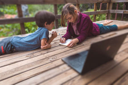 Brother and sister are doing homework while sitting on the veranda of a house, a girl helps her little brother, children are writing in a notebook, a child is looking at a laptop, distance educationの写真素材