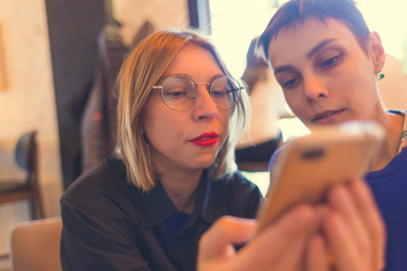 Two girls are sitting in a coffee shop. Woman shows her friend a mobile phone. Blonde with glasses and a brunette with short hair.の写真素材