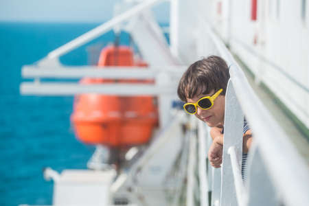 A child is sailing on a cruise ship, a boy in sunglasses stands on board the ship, a ferry is sailing on the sea, a child is traveling and looking at the waves.の写真素材