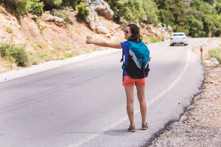 A woman stops a car on the road, travel through scenic places, a girl hitchhikes, a woman with a backpack is waiting for transport on a mountain roadの写真素材