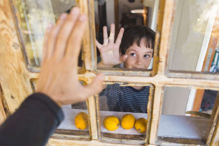 A curious boy looks out the window, Portrait of a boy, Observation through the window, the emotions of a child, kid looks out of the window of a house, quarantine self-isolation, boy waving.の写真素材