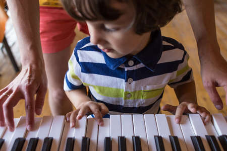 A woman teaches her son to play the piano. The boy masters the keyboard musical instrument. A child learns music. Children's and women's hands on the piano keys. Music teacher. Tutor for a child.の写真素材