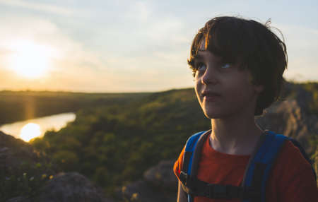 Portrait of a boy with a backpack on the background of the setting sun, boy with a backpack on a hike, a hike with a child, summer vacation in nature.の写真素材