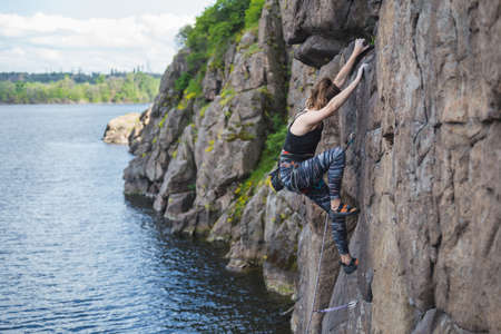 A girl climbs a rock above the water, a woman is engaged in extreme sports, rock climbing on natural terrain, strength and endurance trainingの写真素材