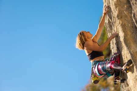 The girl climbs the rock. The climber trains on natural terrain. A woman overcomes a difficult climbing route against the background of a blue sky.の写真素材