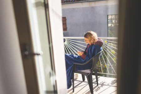 a girl with a phone sits on a sunny balcony, covered with a blanket.の写真素材
