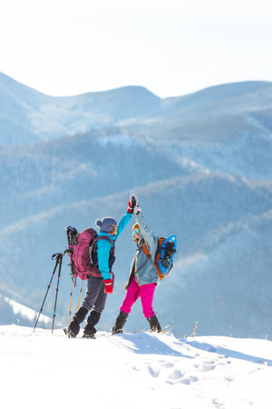 two women climbed to the top of the mountain during a winter hike, winter trekking, two girlfriends travel together, snow-capped mountains, girl gives high five to friendの写真素材