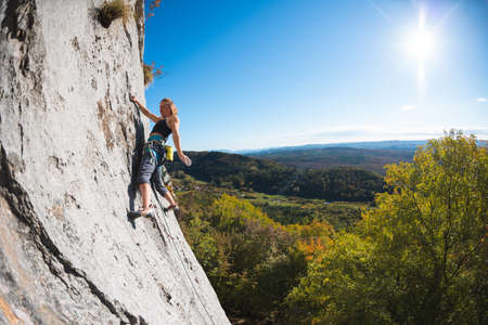 girl rock-climber climbs the rock against the background of the autumn forest and blue sky. a climber walks a difficult route using equipment.の写真素材