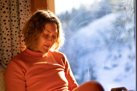 a woman sits near the window and holds a smartphone, a smiling girl near the window on a sunny day, a cozy placeの写真素材