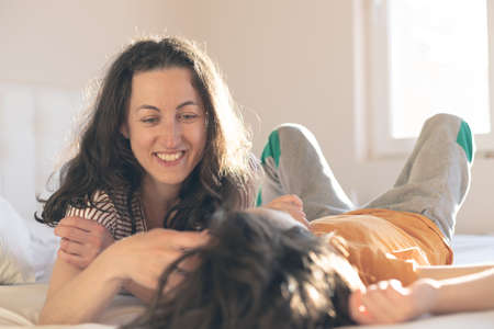 Mother strokes her son on the head, the boy with his mother lies on the bed, the child spends the morning with his mother, the woman touches the child's hair, mother's dayの写真素材