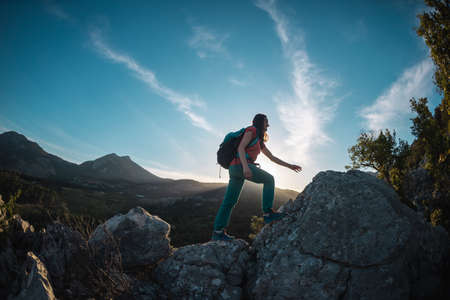 A woman with a backpack climbs to the top, hiking in the mountains, success in achieving the goal, the silhouette of a girl against the backdrop of a mountain at sunsetの写真素材