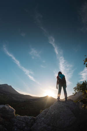 A woman with a backpack climbed to the top and looks into the distance, hiking in the mountains, success in achieving the goal, the silhouette of a girl against the backdrop of a mountain at sunsetの写真素材