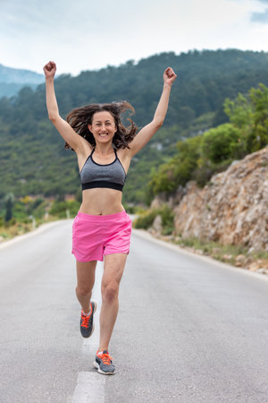 A girl runs along an asphalt road against the backdrop of beautiful mountains, a woman on an evening jog, a slender woman runs along a mountain road, The runner finished and put his hands up, victoryの写真素材