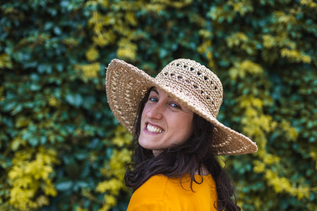 A smiling girl in a straw hat stands near a wall overgrown with ivy, a portrait of a woman with dark...
