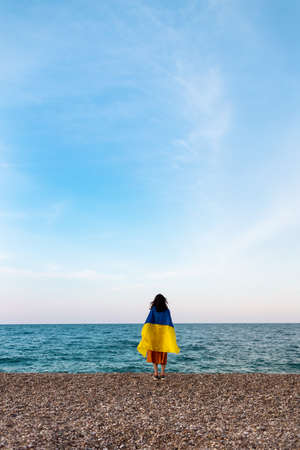 A girl stands by the sea with a Ukrainian flag, support for Ukraine, a sad Ukrainian woman, the flag flutters in the windの写真素材