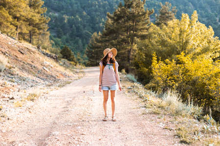 A girl with a backpack and a straw hat against the backdrop of a large mountain, a young woman walks along a mountain path, a trip to the beautiful places of the worldの写真素材