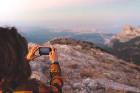 beautiful young girl travels and takes a photo on the phone. travel in the mountains. portrait of a girl with a phone at sunset.の写真素材