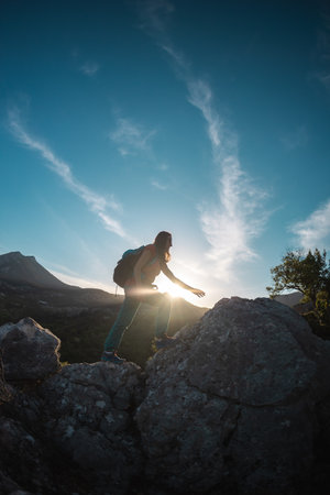 A woman with a backpack climbs to the top, hiking in the mountains, success in achieving the goal, the silhouette of a girl against the backdrop of a mountain at sunsetの写真素材