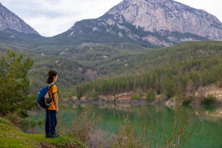 A child with a backpack looks at a beautiful mountain lake. Relaxed, peaceful, thoughtful, happy and free on a mountain lake. vacation with childrenの写真素材
