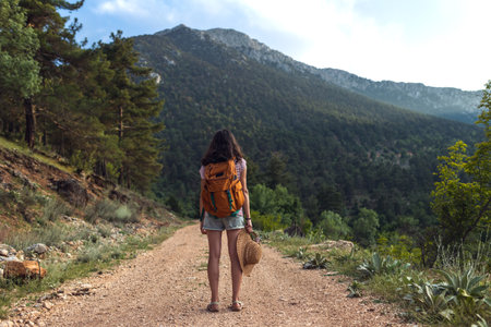 girl with a backpack enjoys the journey. the traveler stands on the road in the forest against the backdrop of mountains. adventure and hiking.の写真素材