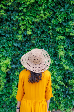 young girl in a hat and yellow dress stands against a wall with green bushes. happy girl on...