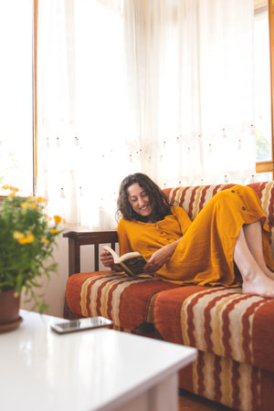 The girl lies on the sofa and reads a book, a young woman in a dress reads at homeの写真素材