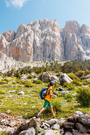 children's trip. child traveler with a backpack resting outdoors with rocky mountains in the background. Concept of summer vacation and hiking lifestyleの写真素材