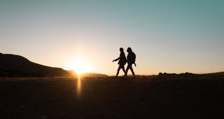 silhouette of two girls with backpacks while traveling in the mountains at sunset. two people walk along the mountain range at sunset. travel with freedom.の写真素材