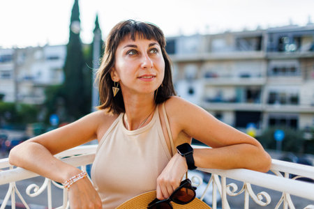 young girl is standing on the balcony. portrait of a beautiful girl with a hat and glasses in her hands on a balcony in the city. vacation in the city.の写真素材