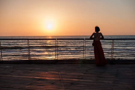 A woman enjoys the sunset while traveling. Elegant happy woman in a dress looks at the ocean.の写真素材