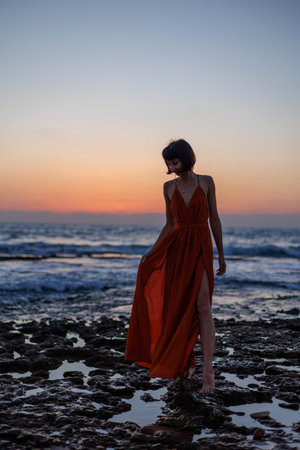 young woman in a summer dress walks along the seashore during sunset and looks at the sea. freedom and beauty.の写真素材