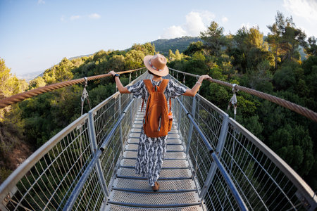happy laughing woman in a hat walks along a pedestrian suspension bridge over the river on a sunny autumn day. back view.の写真素材
