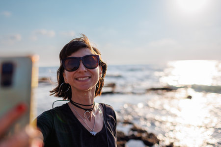Close-up of a stunning woman with hair blowing in the wind taking a selfie by the sea. Beautiful girl smiling while looking at camera outside - fashionable lifestyle and beauty conceptの写真素材