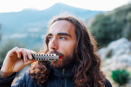 a young man with long hair plays the harmonica. a man enjoys playing the harmonica while standing on the street.の写真素材