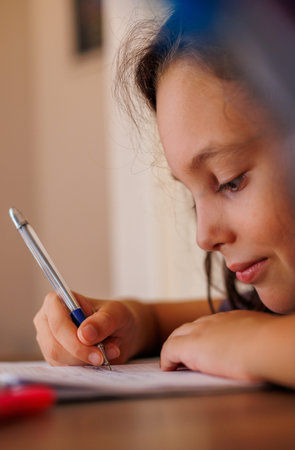 Side view of a focused little boy who is learning to write while doing his homework while sitting at...