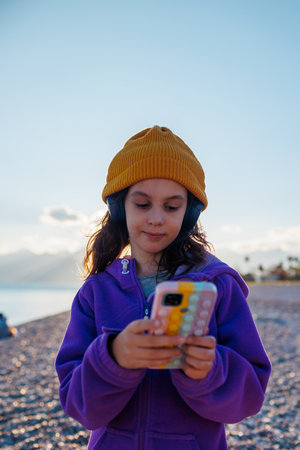child with mobile phone and headphones listens to music, style and fashion. beach and mountains in the backgroundの写真素材