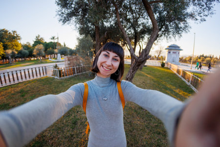Smiling girl takes selfies, video chats, holds smartphone and talks to camera while posing in park....