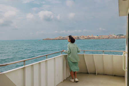 A young woman stands on the deck of a cruise ship and looks at the city. The girl is traveling. travel by ship. View of the city and port in the city of Akko (Acre), Israel. travel and adventure.の写真素材