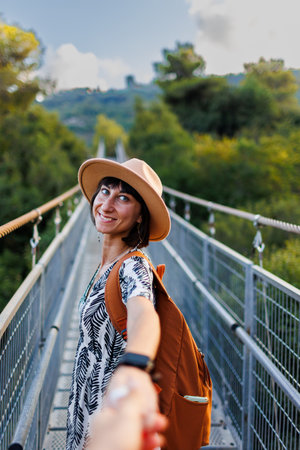Girl with a backpack. outdoor portrait of a young woman walking on a suspension bridge over a river, the girl turns and extends her hand to the camera as if she is following me. Travel and adventure.の写真素材