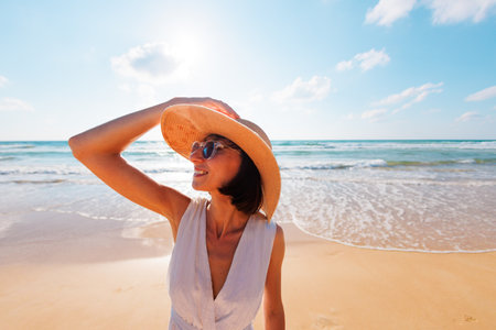 Young joyful woman in a white dress and hat smiling at the camera on the beach. Girl traveler enjoying freedom outdoors on a sunny day. Concept of healthy lifestyle and happy peopleの写真素材