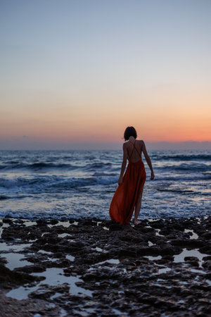 young woman in a summer dress walks along the seashore during sunset and looks at the sea. freedom and beauty.の写真素材