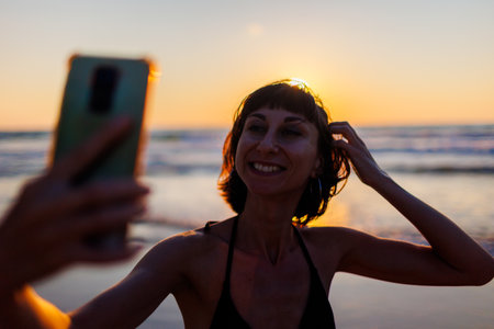 Beautiful teenager with smartphone walking along sea beach at sunset, taking selfie. Girl smiling phone camera standing beach at sunrise closeup. Woman making selfieの写真素材