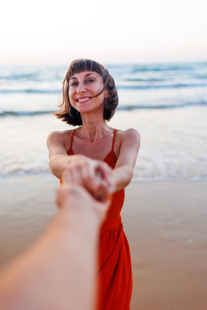 happy young woman pulls guy's hand. girl having fun on the beach. a beautiful girl in a dress walks along the beach, holding a guyの写真素材