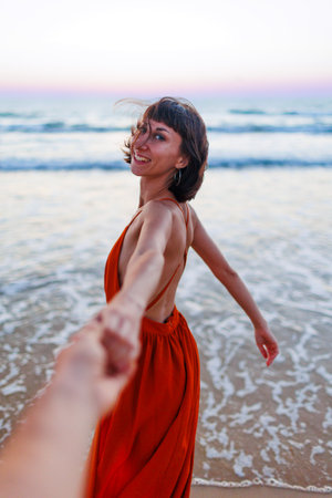 happy young woman pulls guy's hand. girl having fun on the beach. a beautiful girl in a dress walks along the beach, holding a guyの写真素材