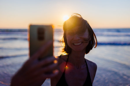 Beautiful teenager with smartphone walking along sea beach at sunset, taking selfie. Girl smiling...