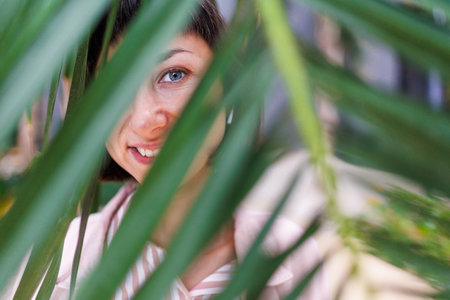 Close-up portrait of a woman standing behind palm leaves. Natural beauty against the backdrop of tropical leaves. Summer girl in a pink shirt behind palm leaves.の写真素材