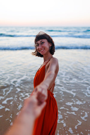 happy young woman pulls guy's hand. girl having fun on the beach. Follow me. a beautiful girl in a dress walks along the beach, holding a guyの写真素材